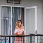 Winsome tanned girl with phone in hands smiling and looking away. Cheerful young lady in pink outfit standing at hotel balcony.